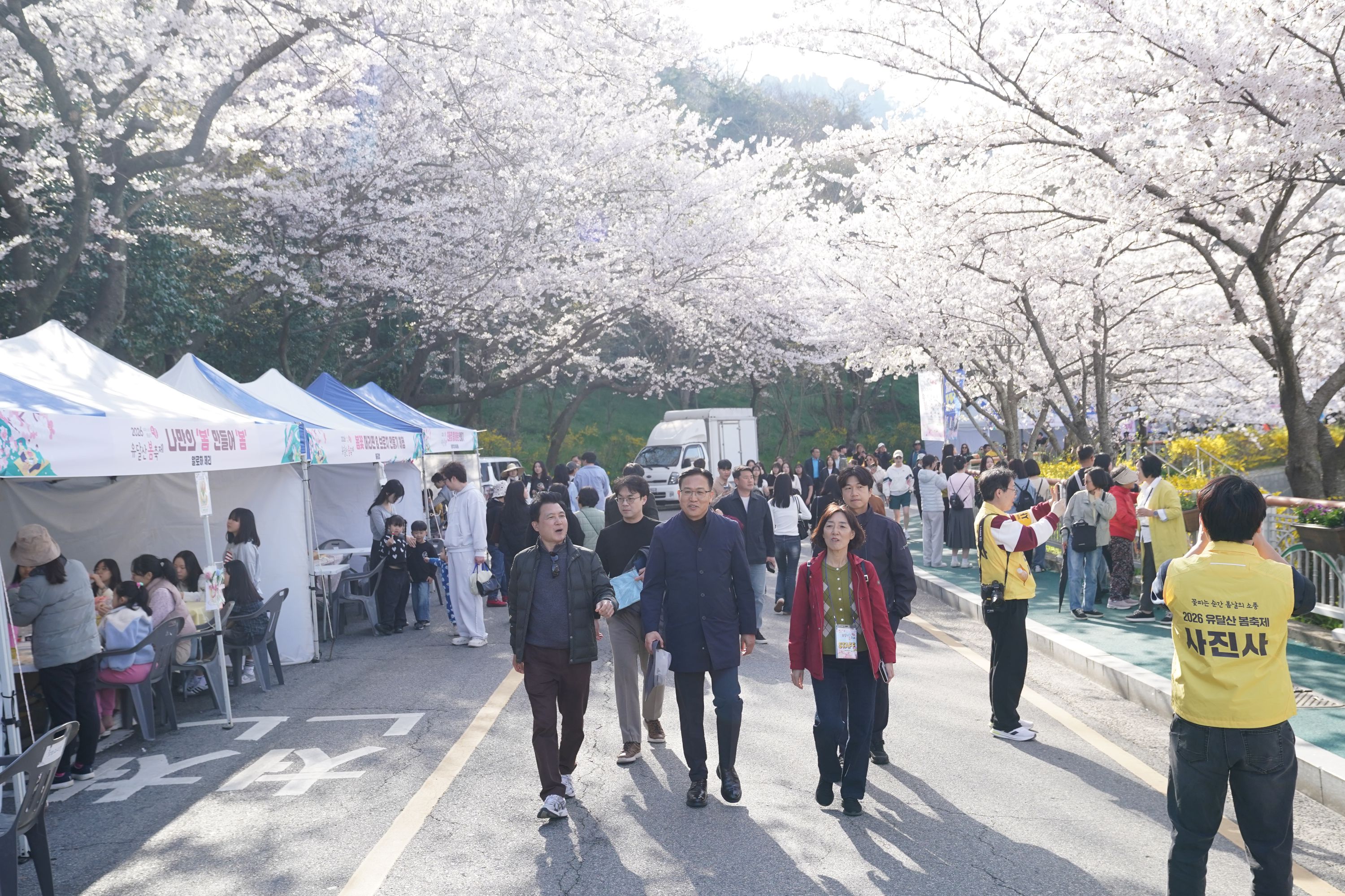 야외 도로에서 많은 사람들이 걸어 다니고 있는 모습이다. 주변에는 벚꽃이 핀 나무가 보이며, 도로 양쪽으로 사람들이 모여 이동하고 있다. 근처 건물과 상점 간판이 보이고, 일부 사람들은 풍선을 들고 있다.