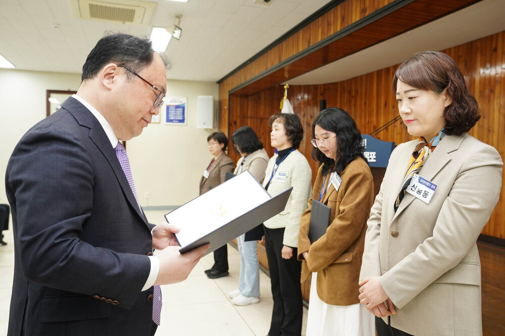 4월 정례조회에서 정장을 입은 남성이 베이지 정장과 스카프를 착용한 여성에게 표창장을 전달하는 모습