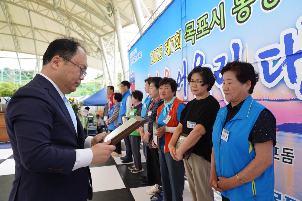 정장을 입고 안경을 낀 남성이 노란색 도트무늬가 그려진 검은색 반팔티에 파란색 작업 조까를 입은 여성에게 패를 수여하고 있다