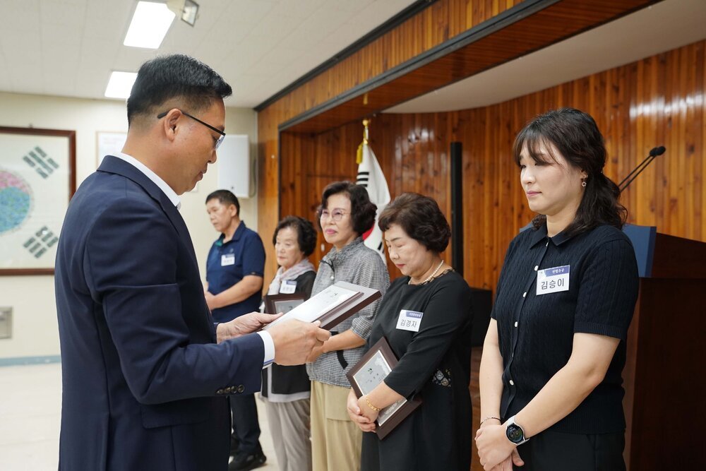 정장 차림의 관계자가 검은색 반팔 니트를 입은 여성 수상자에게 패를 전달하며 읽고 있는 모습이다.