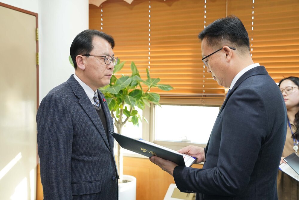 2026년 상반기 4급이하 정기인사로 정장을 입은 남성이 실내 사무실에서 한 남성에게 상장을 전달하며 내용을 읽고 있고, 남성은 안경을 쓰고 짙은 회색 자켓을 입은 채 두 손을 내린 상태로 서 있으며 뒤쪽으로는 블라인드가 내려진 창문과 화분이 보인다.