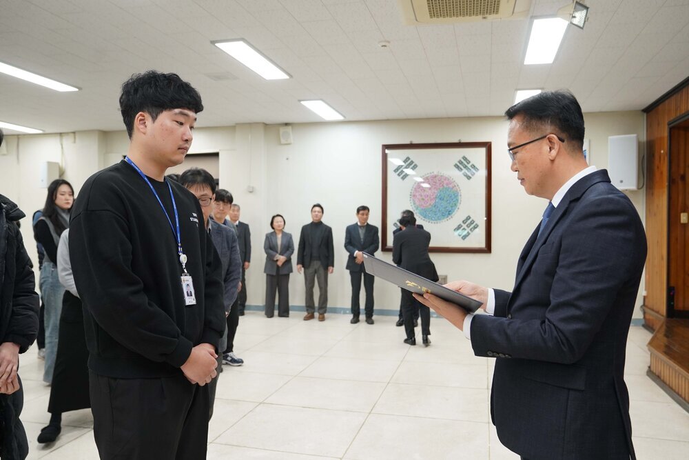 2026년 상반기 4급이하 정기인사로 정장을 입은 남성이 실내 행사장에서 한 남성에게 상장을 전달하며 내용을 읽고 있고, 남성은 검은 맨투맨을 입은 채 두 손을 모은 상태로 서 있으며 뒤쪽에는 참석자들이 줄지어 서서 행사를 지켜보고 있다.