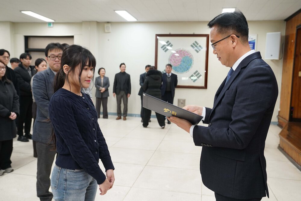 2026년 상반기 4급이하 정기인사로 정장을 입고 안경을 쓴 남성이 실내 행사장에서 한 여성에게 상장을 전달하며 내용을 읽고 있고, 여성은 남색 니트를 입고 두 손을 모은 채 서 있으며 뒤쪽에는 참석자들이 줄지어 서서 행사를 지켜보고 있다.