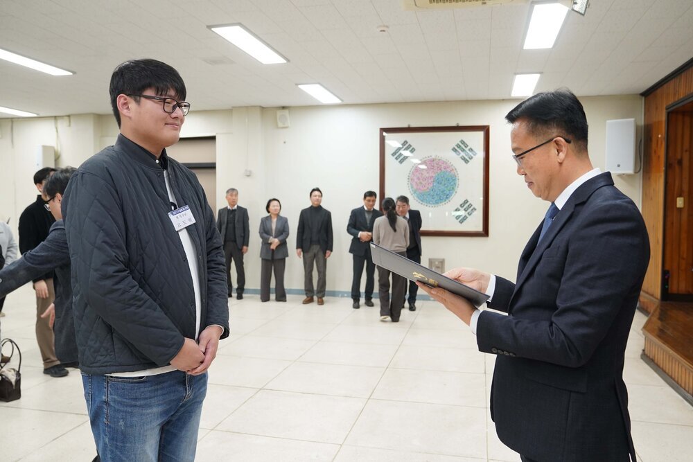 2026년 상반기 4급이하 정기인사로 정장을 입고 안경을 쓴 남성이 실내 행사장에서 한 남성에게 상장을 전달하며 내용을 읽고 있고, 남성은 검은 자켓과 청바지를 입고 서 있으며 뒤쪽에는 참석자들이 줄지어 서서 행사를 지켜보고 있다.