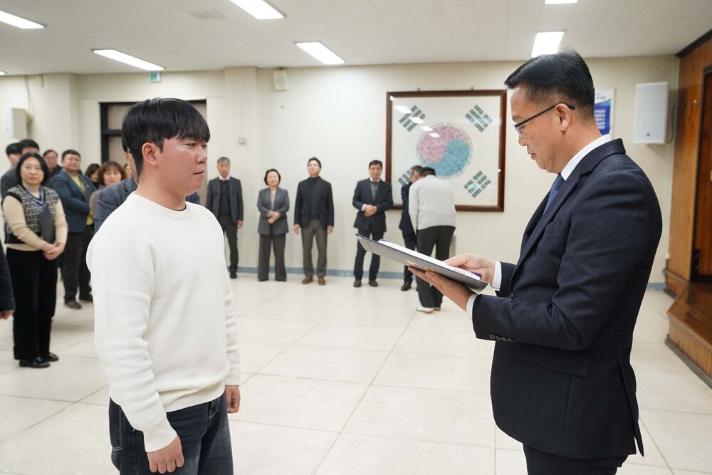 2026년 상반기 4급이하 정기인사로 정장을 입고 안경을 쓴 남성이 실내 행사장에서 한 남성에게 상장을 전달하며 내용을 읽고 있고, 남성은 흰색 니트를 입고 서 있으며 뒤쪽에는 여러 사람들이 줄지어 서서 행사를 지켜보고 있다.