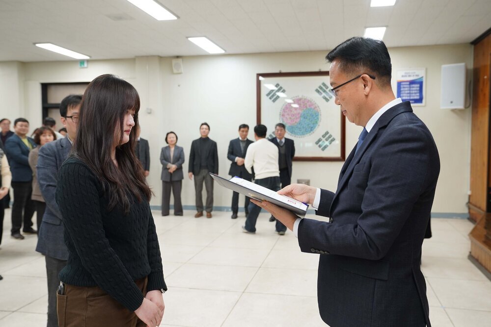 2026년 상반기 4급이하 정기인사로 정장을 입고 안경을 쓴 남성이 실내 행사장에서 한 여성에게 상장을 전달하며 내용을 읽고 있고, 여성은 검은 니트를 입고 두 손을 모은 채 서 있으며 뒤쪽에는 여러 사람들이 줄지어 서서 행사를 지켜보고 있다.