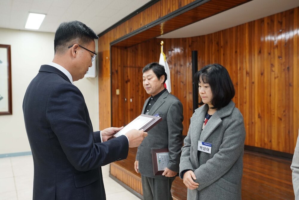 2월 정례조회에 정장을 입고 안경을 낀 남성이 회색 코트를 입은 박성아에게 패를 수여하고 있다.