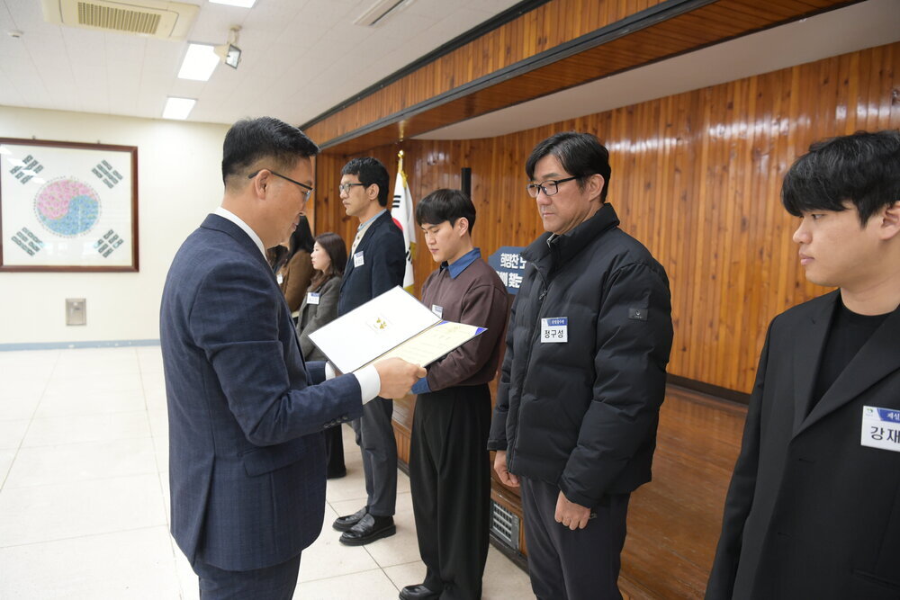 3월 정례조회에 정장을 입고 안경을 낀 남성이 검은바지에 검정색 짧은 패딩을 입은 정구성에게 패를 수여하고 있다.