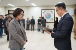 2026년 상반기 4급이하 정기인사로 정장을 입은 남성이 실내 행사장에서 한 여성에게 상장을 전달하며 내용을 읽고 있고, 여성은 짧은 파마머리에 회색 코트를 입고 두 손을 모은 상태로 서 있으며 뒤쪽으로 참석자들이 줄지어 서서 행사를 지켜보고 있다.
