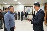 2026년 상반기 4급이하 정기인사로 정장을 입은 남성이 실내 행사장에서 한 남성에게 상장을 전달하며 내용을 읽고 있고, 남성은 안경을 쓰고 하늘색 반집업 니트를 입고 사원증을 착용한 채 두 손을 내린 상태로 서 있으며 뒤쪽으로 참석자들이 줄지어 서서 행사를 지켜보고 있다.