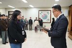 2026년 상반기 4급이하 정기인사로 정장을 입은 남성이 실내 행사장에서 한 여성에게 상장을 전달하며 내용을 읽고 있고, 여성은 안경을 쓰고 검은색 코트와 청바지를 입고 사원증을 착용한 채 두 손을 모은 상태로 서 있으며 뒤쪽으로 참석자들이 줄지어 서서 행사를 지켜보고 있다.