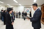 2026년 상반기 4급이하 정기인사로 정장을 입은 남성이 실내 행사장에서 한 여성에게 상장을 전달하며 내용을 읽고 있고, 여성은 검은색 자켓에 초록색 무늬가 있는 스카프를 매고 사원증을 착용한 채 두 손을 모은 상태로 서 있으며 뒤쪽에는 참석자들이 행사를 지켜보고 있다.
