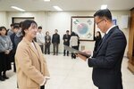 2026년 상반기 4급이하 정기인사로 정장을 입은 남성이 실내 행사장에서 한 여성에게 상장을 전달하며 내용을 읽고 있고, 여성은 체크무늬 머리끈으로 머리를 묶고 밝은 베이지색 코트를 입고 사원증을 착용한 채 두 손을 모은 상태로 서 있으며 뒤쪽으로 참석자들이 줄지어 서서 행사를 지켜보고 있다.