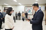 2026년 상반기 4급이하 정기인사로 정장을 입은 남성이 실내 행사장에서 한 여성에게 상장을 전달하며 내용을 읽고 있고, 여성은 흰색 트위드 자켓을 입고 두 손을 모은 상태로 서 있으며 뒤쪽으로 참석자들이 줄지어 서서 행사를 지켜보고 있다.