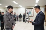 2026년 상반기 4급이하 정기인사로 정장을 입은 남성이 실내 행사장에서 한 남성에게 상장을 전달하며 내용을 읽고 있고, 남성은 회색 집업 점퍼를 입고 사원증을 착용한 채 두 손을 모은 상태로 서 있으며 뒤쪽으로 참석자들이 줄지어 서서 행사를 지켜보고 있다.