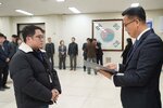 2026년 상반기 4급이하 정기인사로 정장을 입은 남성이 실내 행사장에서 한 남성에게 상장을 전달하며 내용을 읽고 있고, 남성은 안경을 쓰고 목 부분에 흰색 털이 있는 검은색 패딩을 입고 두 손을 모은 상태로 서 있으며 뒤쪽으로 참석자들이 줄지어 서서 행사를 지켜보고 있다.