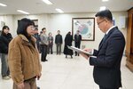 2026년 상반기 4급이하 정기인사로 정장을 입고 안경을 쓴 남성이 실내 행사장에서 한 여성에게 상장을 전달하며 내용을 읽고 있고, 여성은 갈색 퍼 재킷을 입고 안경을 착용한 채 두 손을 모은 채 서 있으며 뒤쪽에는 참석자들이 줄지어 서서 행사를 지켜보고 있다.