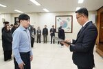 2026년 상반기 4급이하 정기인사로 정장을 입고 안경을 쓴 남성이 실내 행사장에서 한 남성에게 상장을 전달하며 내용을 읽고 있고, 남성은 파란 셔츠를 입고 서 있으며 뒤쪽에는 참석자들이 줄지어 서서 행사를 지켜보고 있다.