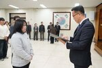 2026년 상반기 4급이하 정기인사로 정장을 입고 안경을 쓴 남성이 실내 행사장에서 한 여성에게 상장을 전달하며 내용을 읽고 있고, 여성은 회색 맨투맨을 입고 두 손을 모은 채 서 있으며 뒤쪽에는 여러 사람들이 줄지어 서서 행사를 지켜보고 있다.