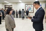 2026년 상반기 4급이하 정기인사로 정장을 입고 안경을 쓴 남성이 실내 행사장에서 한 여성에게 상장을 전달하며 내용을 읽고 있고, 여성은 체크무늬 자켓을 입고 두 손을 모은 채 서 있으며 뒤쪽에는 여러 사람들이 줄지어 서서 행사를 지켜보고 있다.